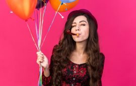 confident young party girl wearing party hat holding balloons blowing party horn with closed eyes isolated on pink background